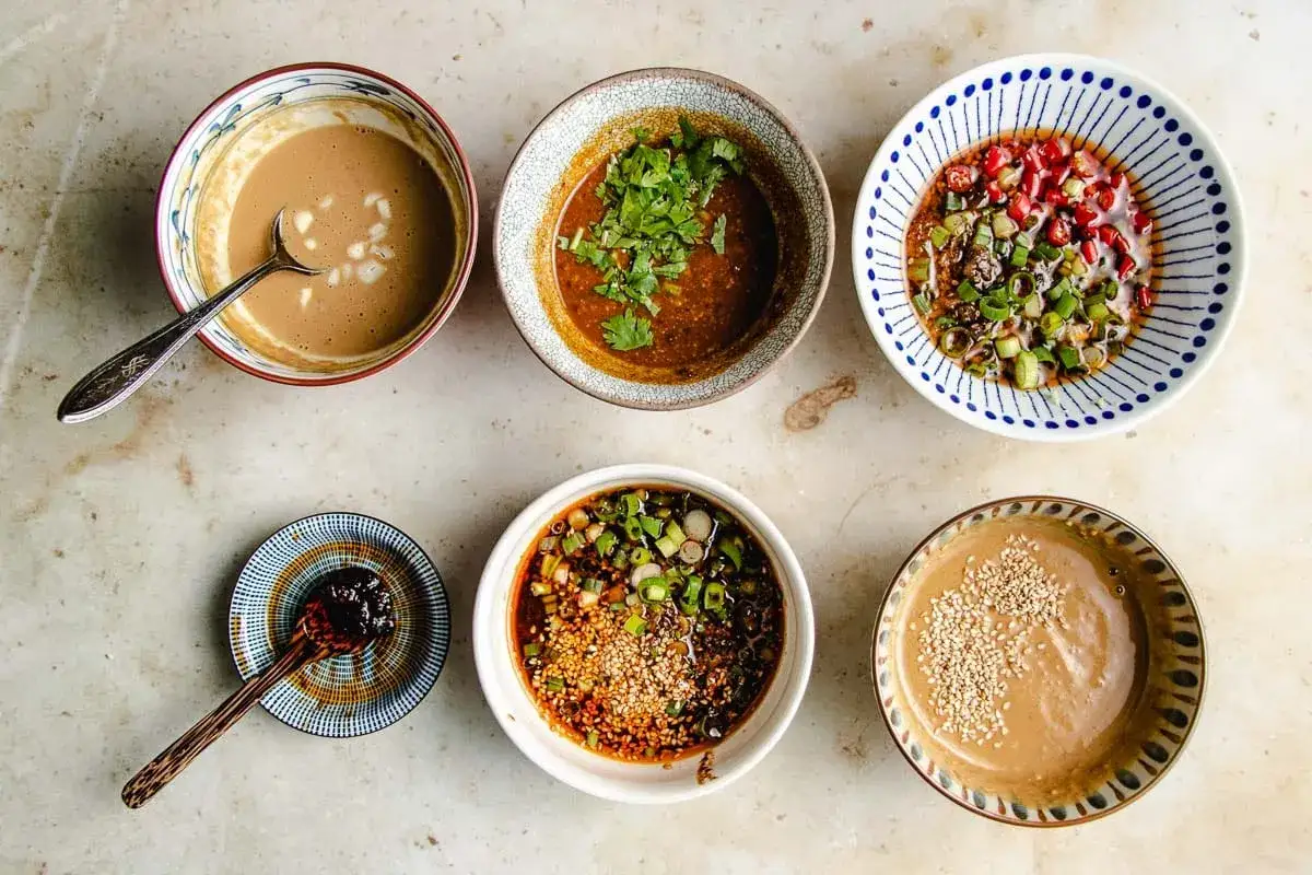 A variety of dipping sauces for hot pot, including creamy sesame sauce, chili oil with scallions, and a spicy pomegranate mixture.