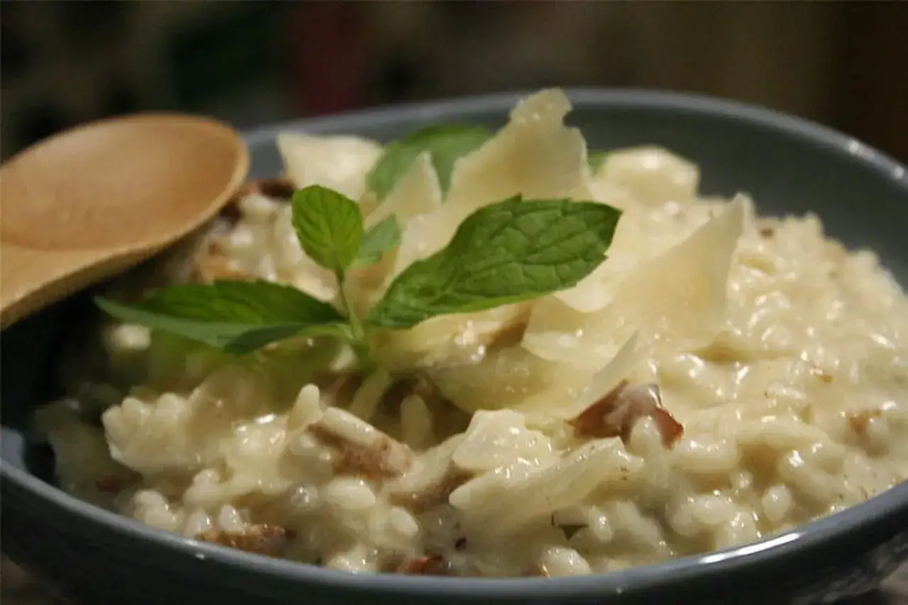 Un crémeux risotto aux champignons, garni de copeaux de parmesan et de feuilles de menthe fraîche. Une cuillère en bois est posée à côté.