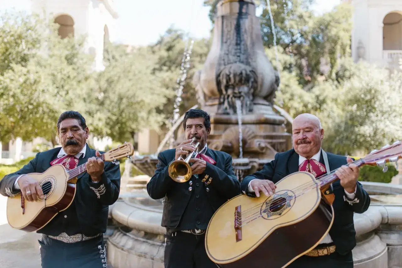 Trzech muzyków mariachi w tradycyjnych strojach gra na gitarach i trąbce przy fontannie.