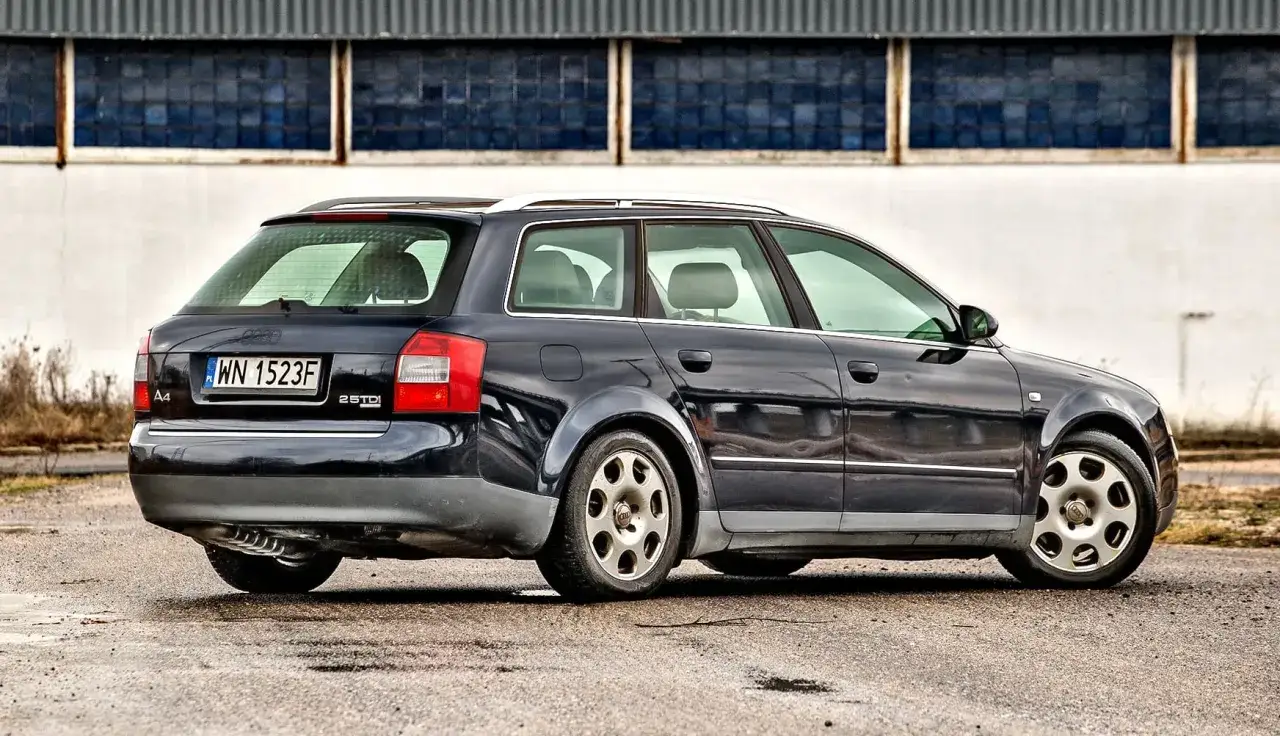 Ciemnogranatowe Audi A4 B6 Avant z polskimi tablicami WN 1523F stoi na mokrym asfalcie przed budynkiem z niebieskimi oknami.