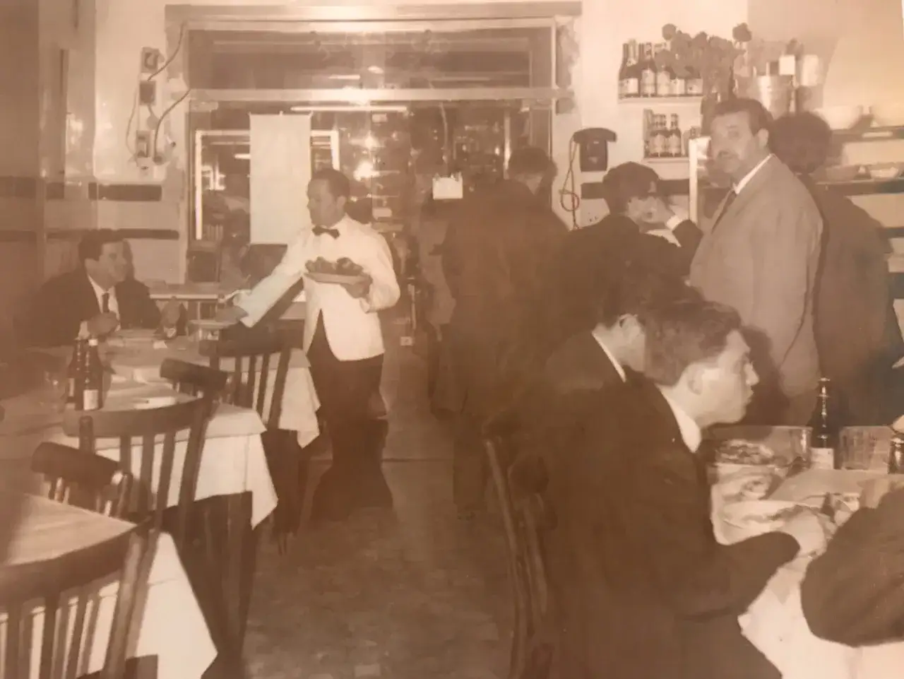 Wnętrze najstarszej pizzerii w Neapolu. Kelner w białej koszuli i muszce niesie talerz z jedzeniem. Przy stolikach siedzą goście.