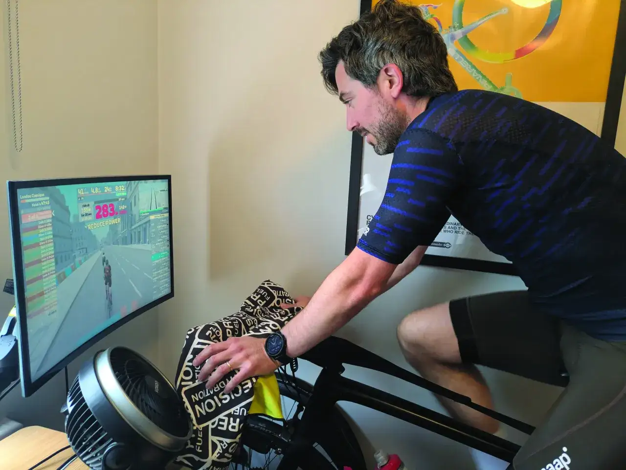 Man doing an indoor sprint workout on a bike trainer, with a fan and a computer screen showing a virtual cycling race.