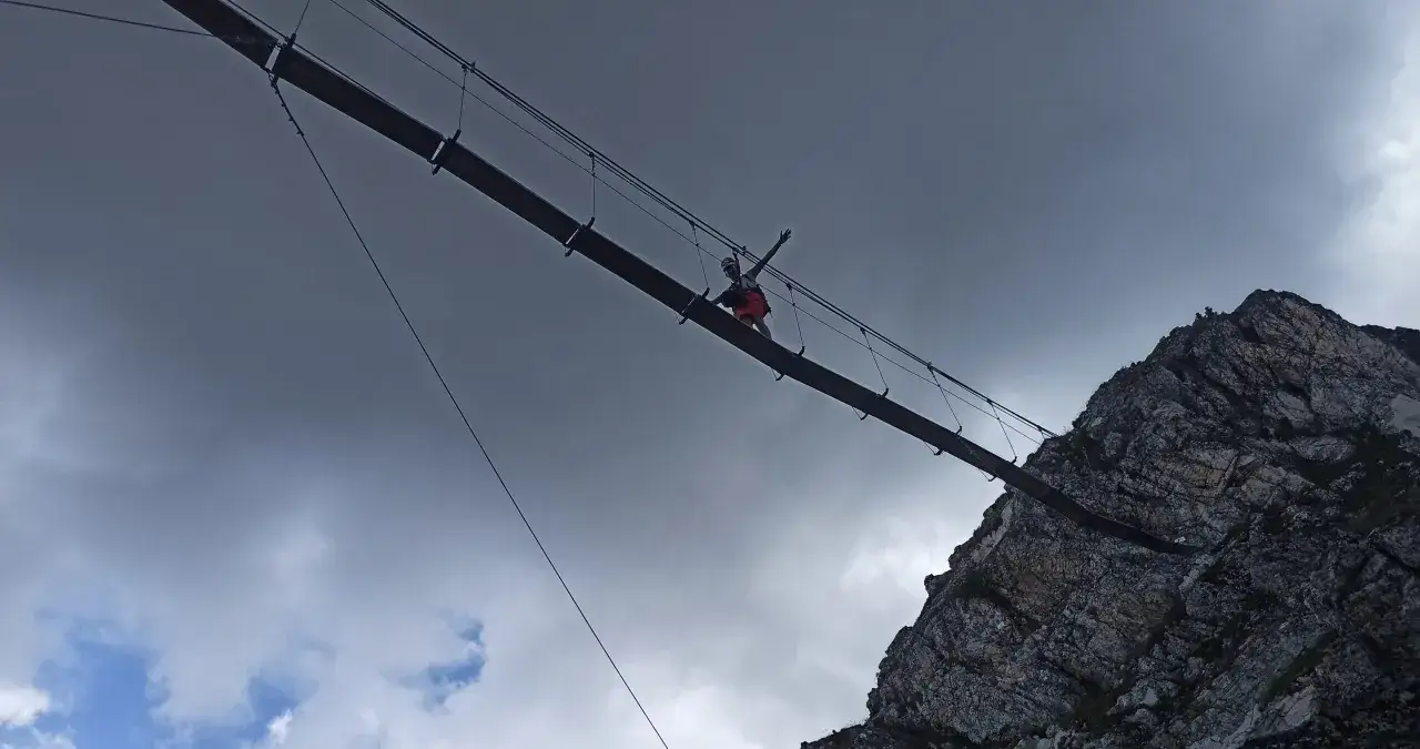 Osoba na moście wiszącym, pokonująca trudną trasę via ferrata polska. W tle skaliste zbocze i burzowe chmury.