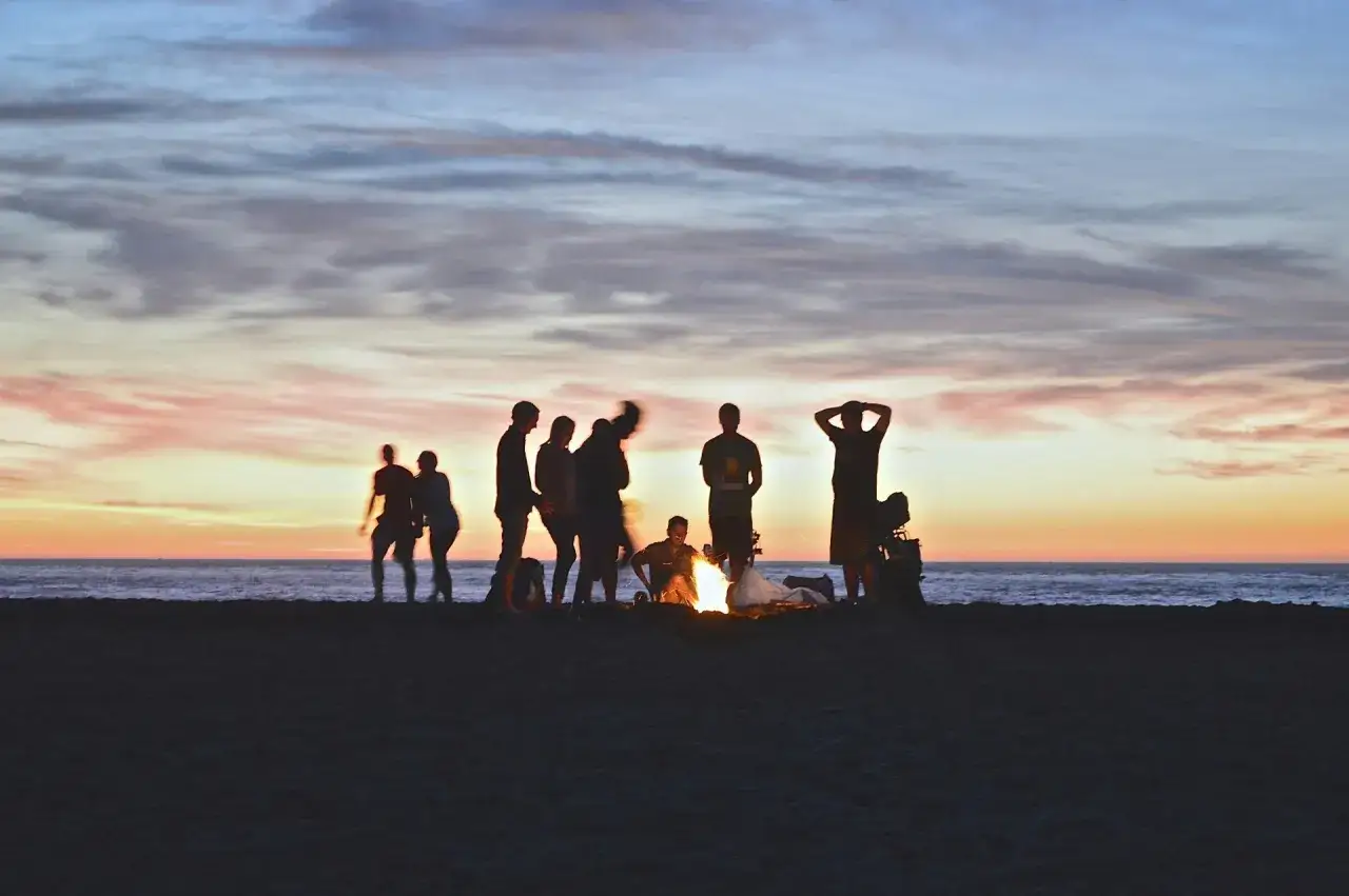 Grupa ludzi przy ognisku na plaży o zachodzie słońca. Czy można spać na plaży? Tak, to idealne miejsce na nocleg.