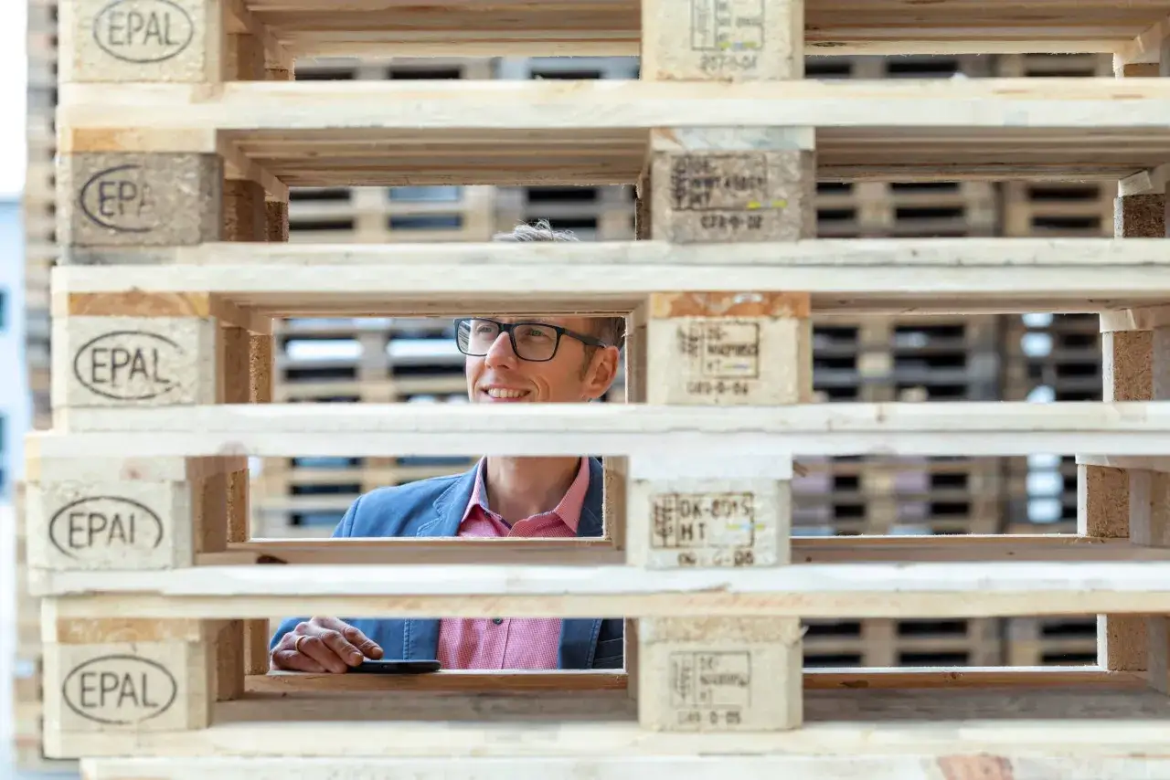 Mann mit Brille l&auml;chelt durch Holzpaletten. EPAL-Siegel auf den Paletten deuten auf einen effizienten Pallet Exchange hin.