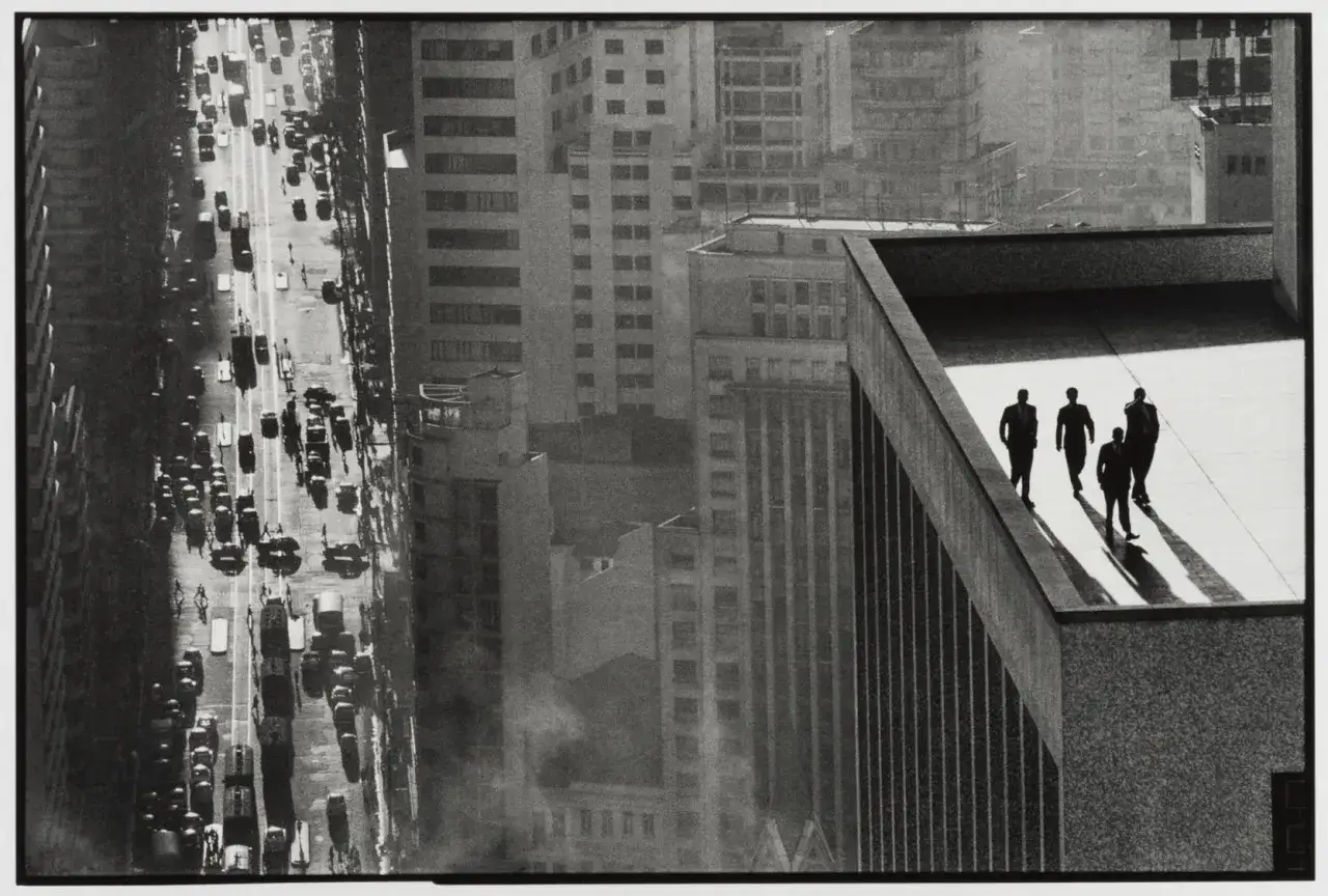 Uomini d'affari camminano sul tetto di un grattacielo, con una strada trafficata sotto. Un'iconica prospettiva di René Burri.
