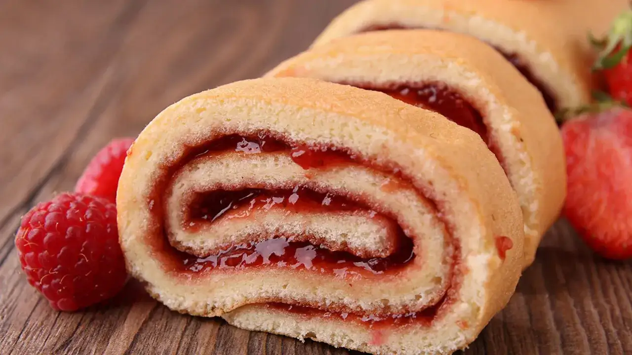 Rebanadas de brazo de gitano con mermelada de fresa y frambuesas frescas sobre una mesa de madera.