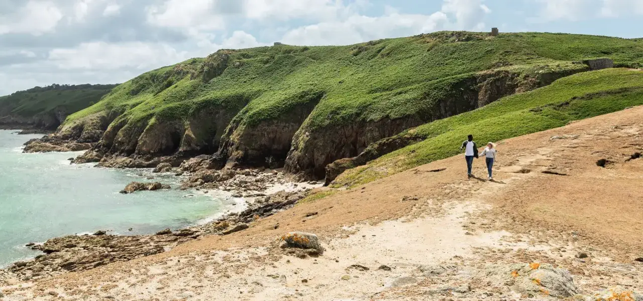 Couple se promenant sur un sentier côtier verdoyant de l'île de Jersey, avec des falaises rocheuses et une mer turquoise.