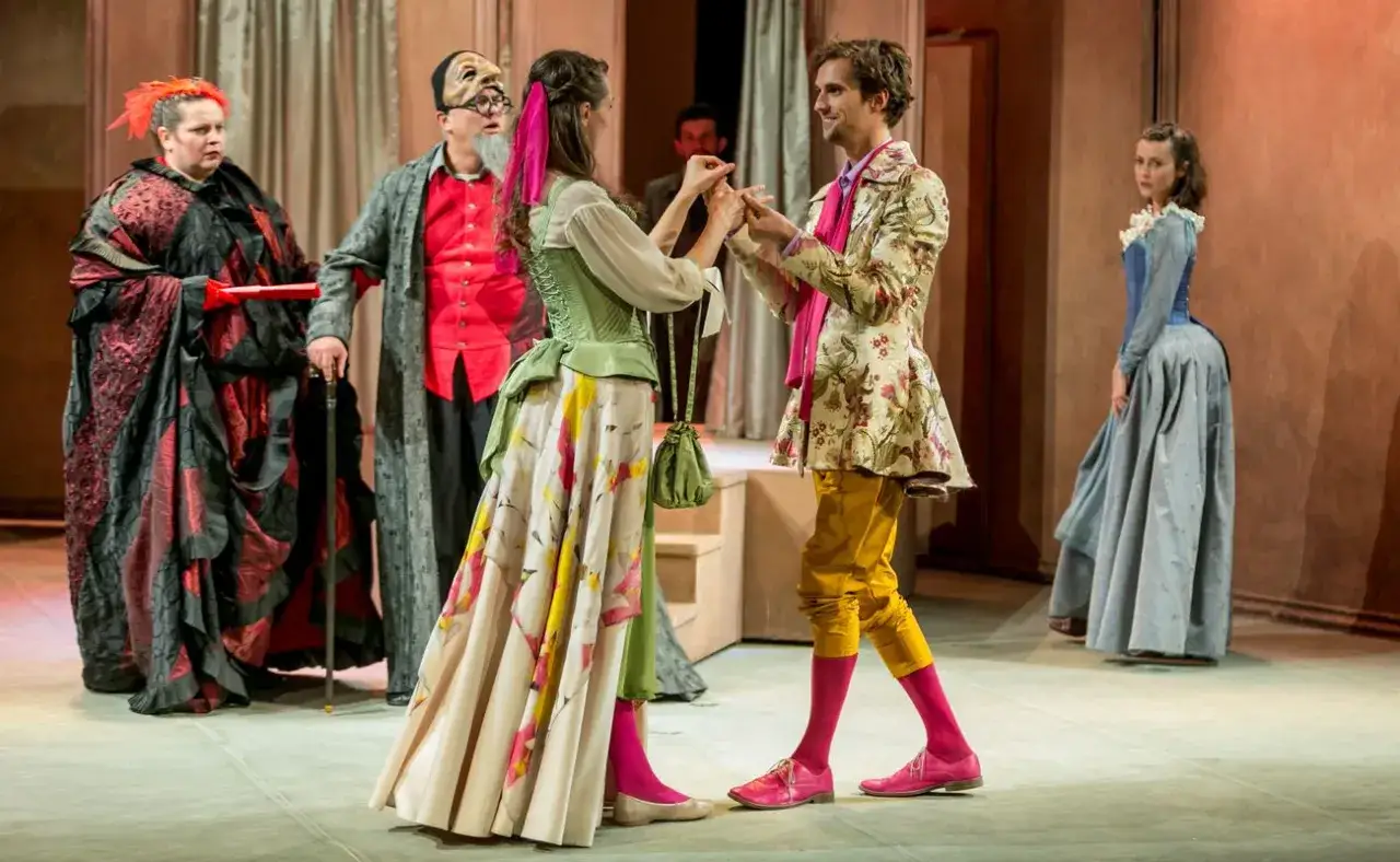 Scena teatralna z epoki, gdzie skąpiec w masce i bogato odziana dama obserwują parę kochanków.