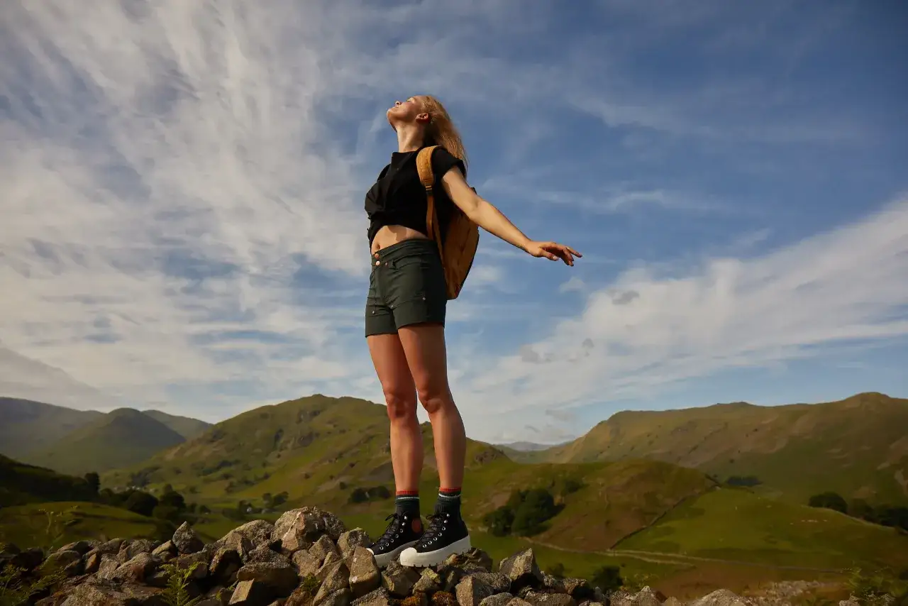 Une jeune femme, sac à dos jaune, short et t-shirt noir, profite du paysage. Idéal pour savoir comment s'habiller pour une randonnée en été.