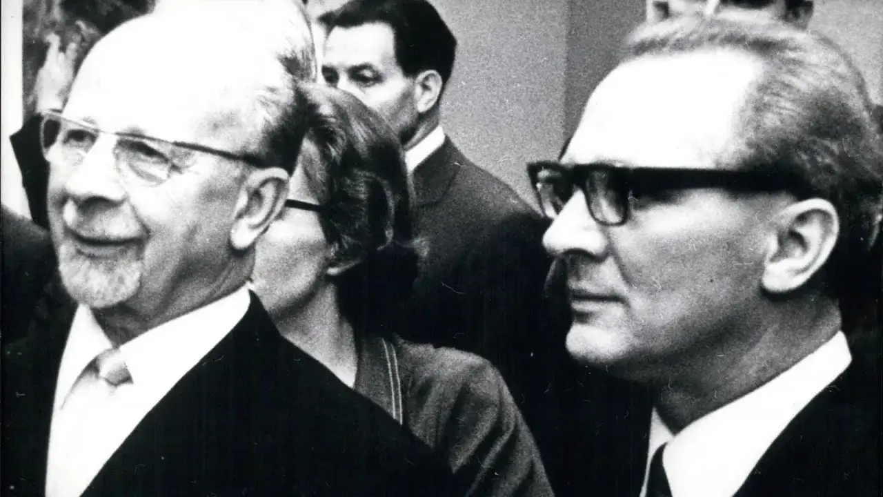 Zwei Männer mit Brillen, einer mit Bart, im Gespräch. Der Mann links könnte ein DDR Chef sein.