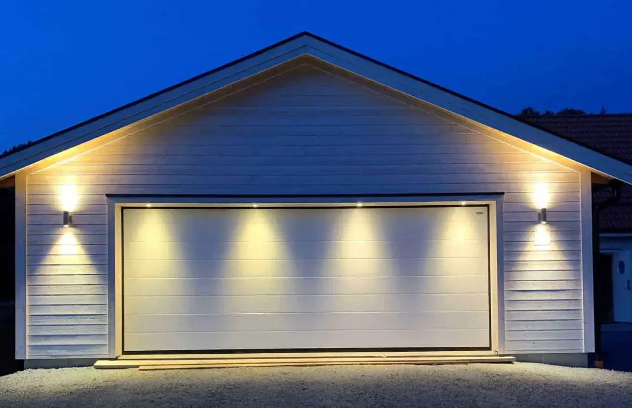 Ett modernt garage med vit port, belyst av utomhusbelysning. En larmskylt utan larm syns inte, men huset ser tryggt ut.