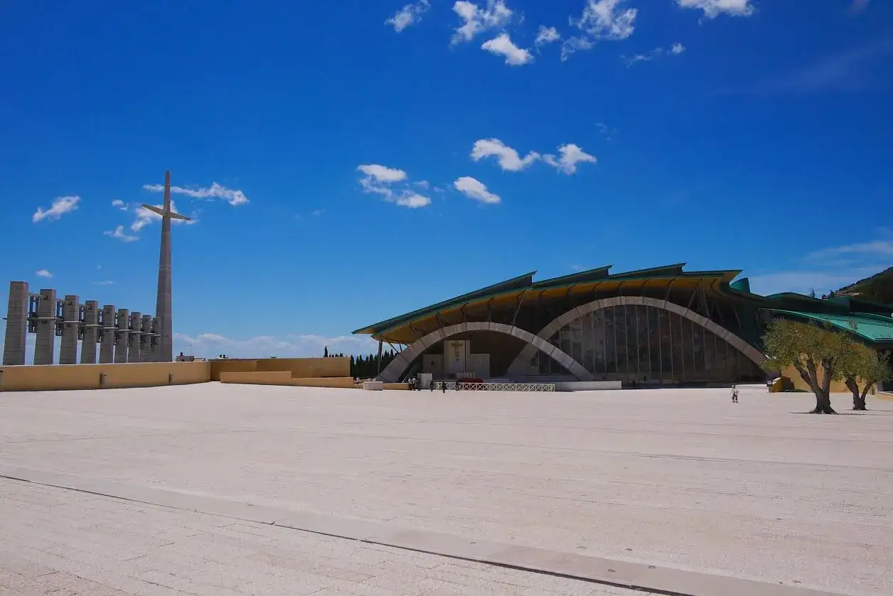 The Sanctuary of Saint Pio of Pietrelcina in San Giovanni Rotondo, Italy, under a bright blue sky. A large cross stands tall, with a modern church building featuring arched windows.