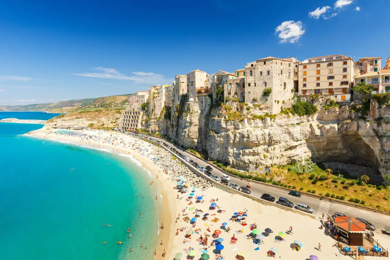 A vibrant beach scene on the Calabria coast, with turquoise waters, a sandy shore dotted with sunbathers, and colorful buildings perched atop dramatic cliffs.
