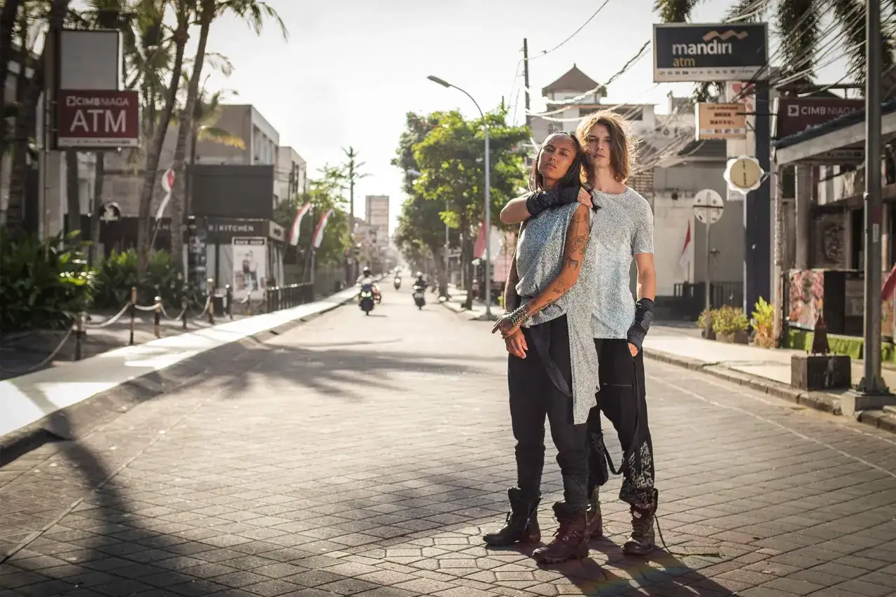 Two individuals showcase gender fluid fashion on a sun-drenched street, their styles blending comfort and edgy details.
