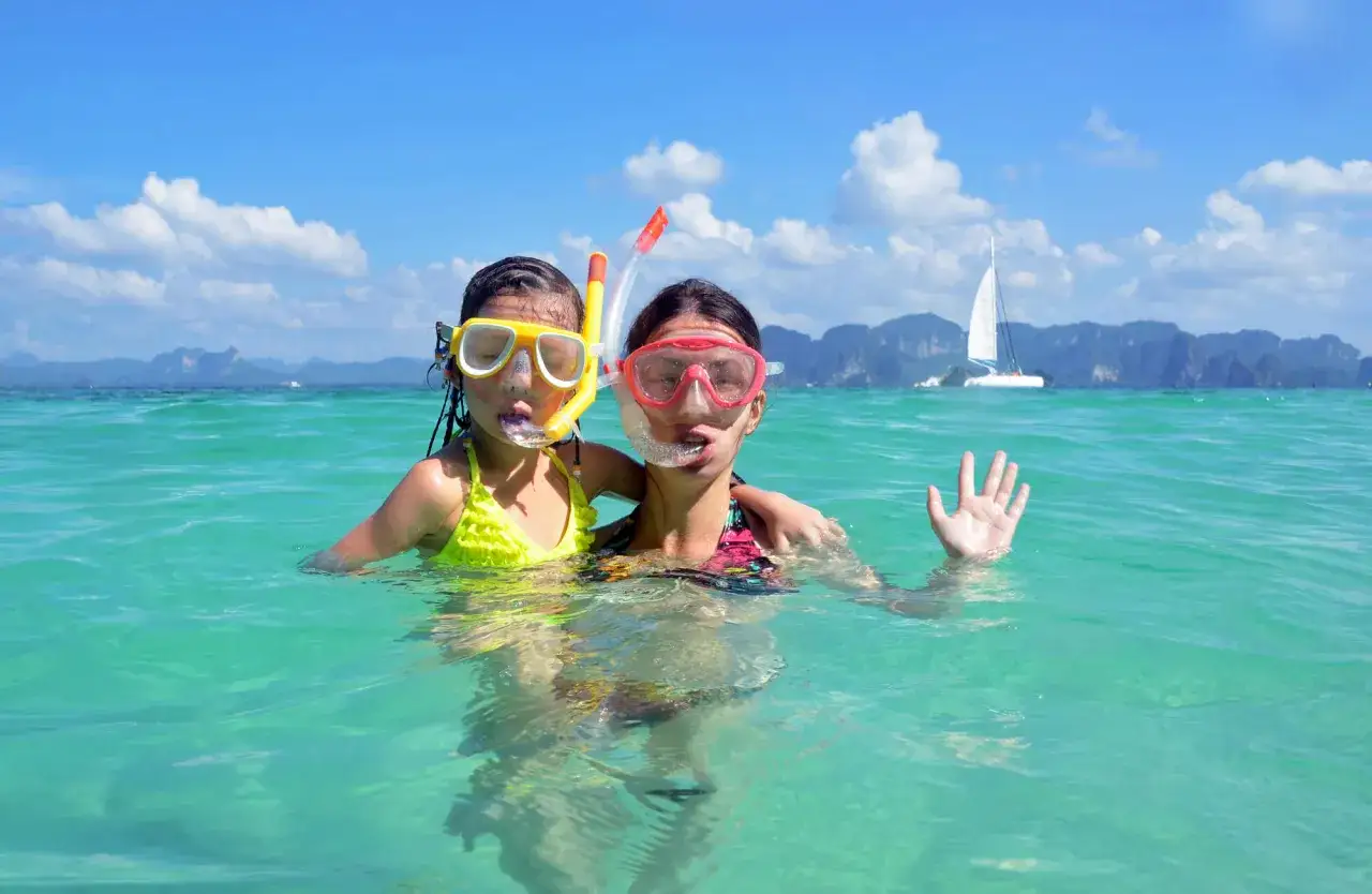 Due bambine con maschere da snorkeling si divertono nell'acqua cristallina della Thailandia, con una barca a vela sullo sfondo.