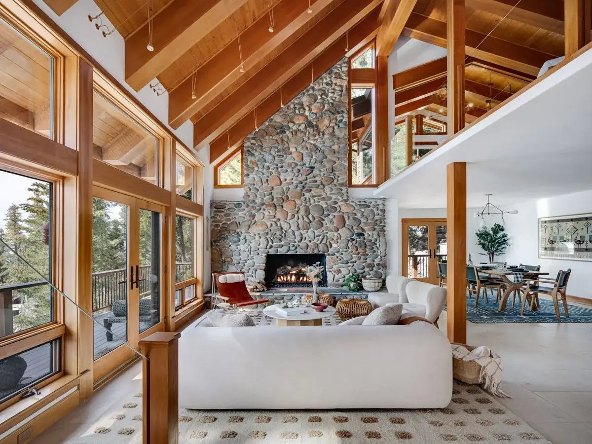Amplio salón con chimenea de piedra, vigas de madera y grandes ventanales. Un sofá blanco y una silla roja crean un ambiente acogedor de estilo rústico.