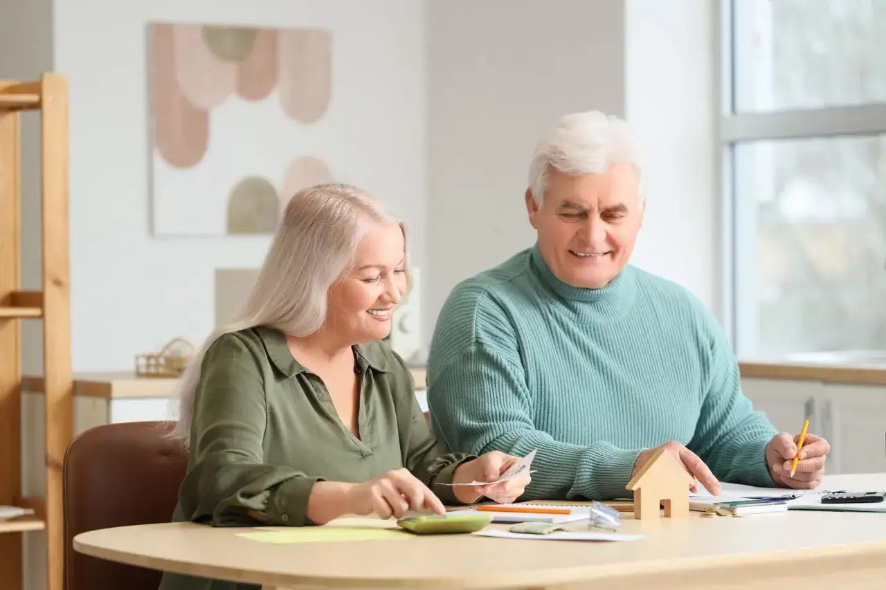 Couple âgé planifiant leur budget pour l'entrée en EHPAD et la gestion de leur résidence principale. Ils sourient en examinant des papiers et une calculatrice.