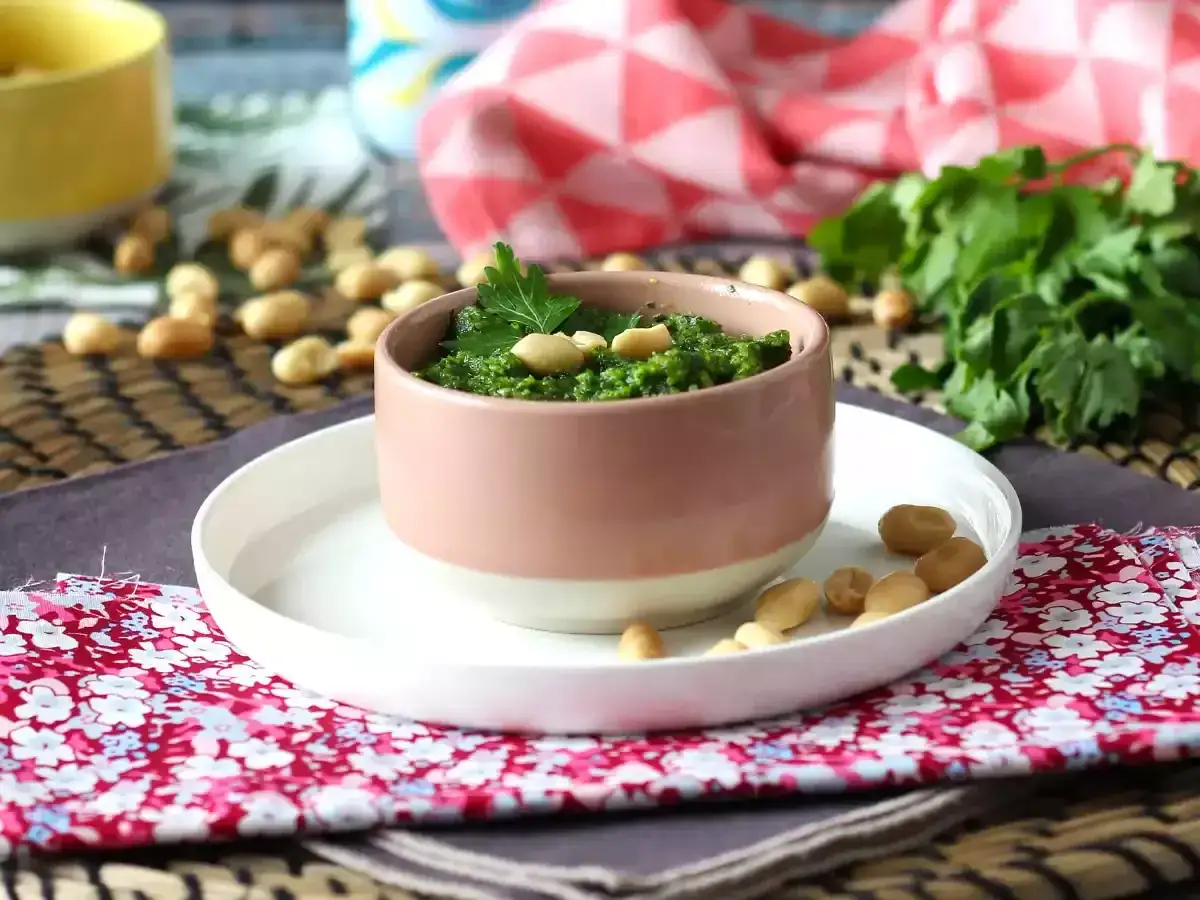 Pesto z pietruszki w różowej miseczce, ozdobione listkiem i orzeszkami. Obok świeża natka pietruszki i orzechy.