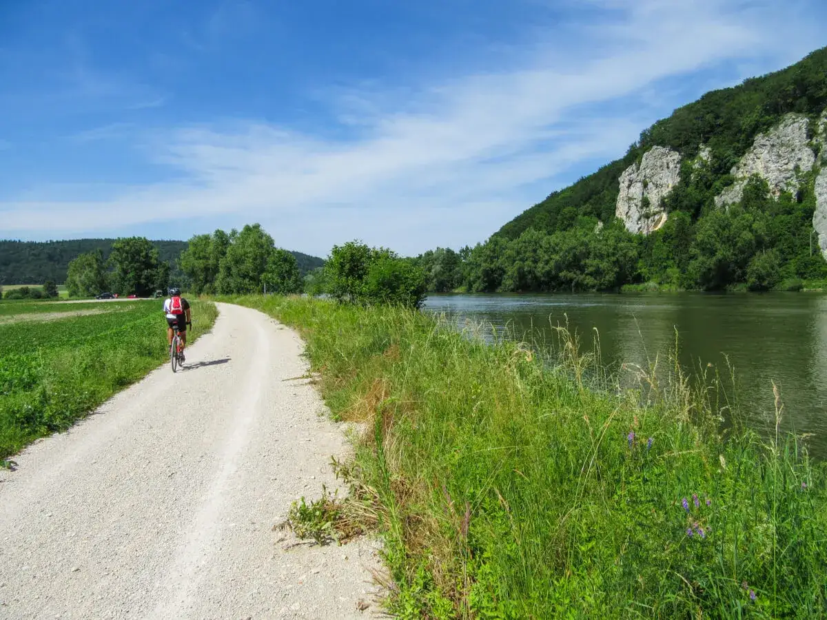 A cyclist enjoys a scenic Danube bike tour along a gravel path beside a wide river and lush green hills.