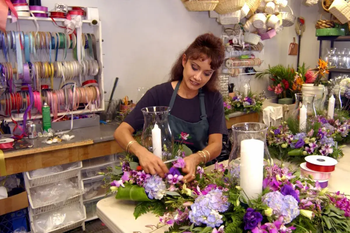 Florysta kto to? Kobieta tworzy bukiet z fioletowych kwiatów i świec. W tle widać rolki wstążek i kosze.