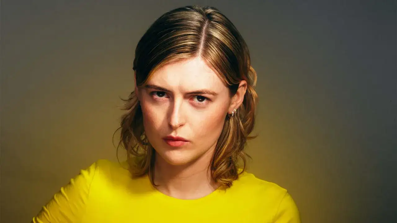 Ellen Krauss, med sitt blonda hår och gula tröja, ser intensivt in i kameran. En bild som fångar energin inför en kommande ellen krauss konsert.