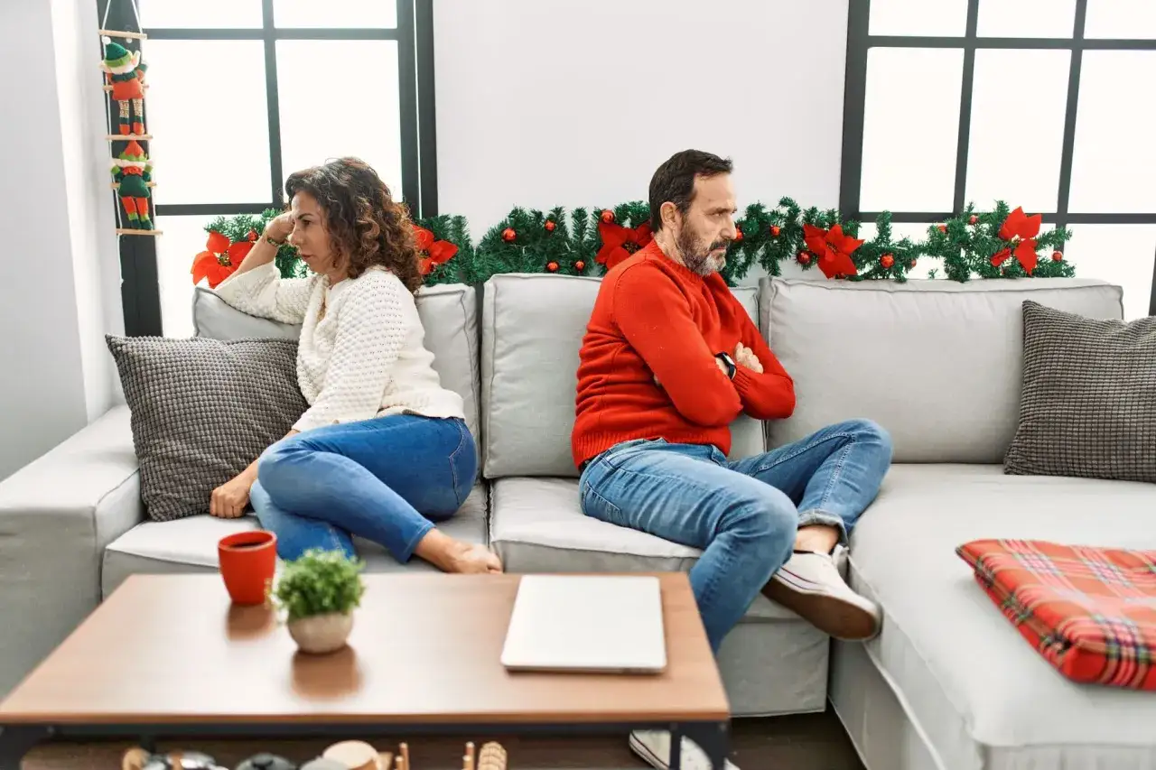 Un couple, visiblement en d&eacute;saccord familial au sujet d'une maison de retraite, est assis dos &agrave; dos sur un canap&eacute;, l'air contrari&eacute;.