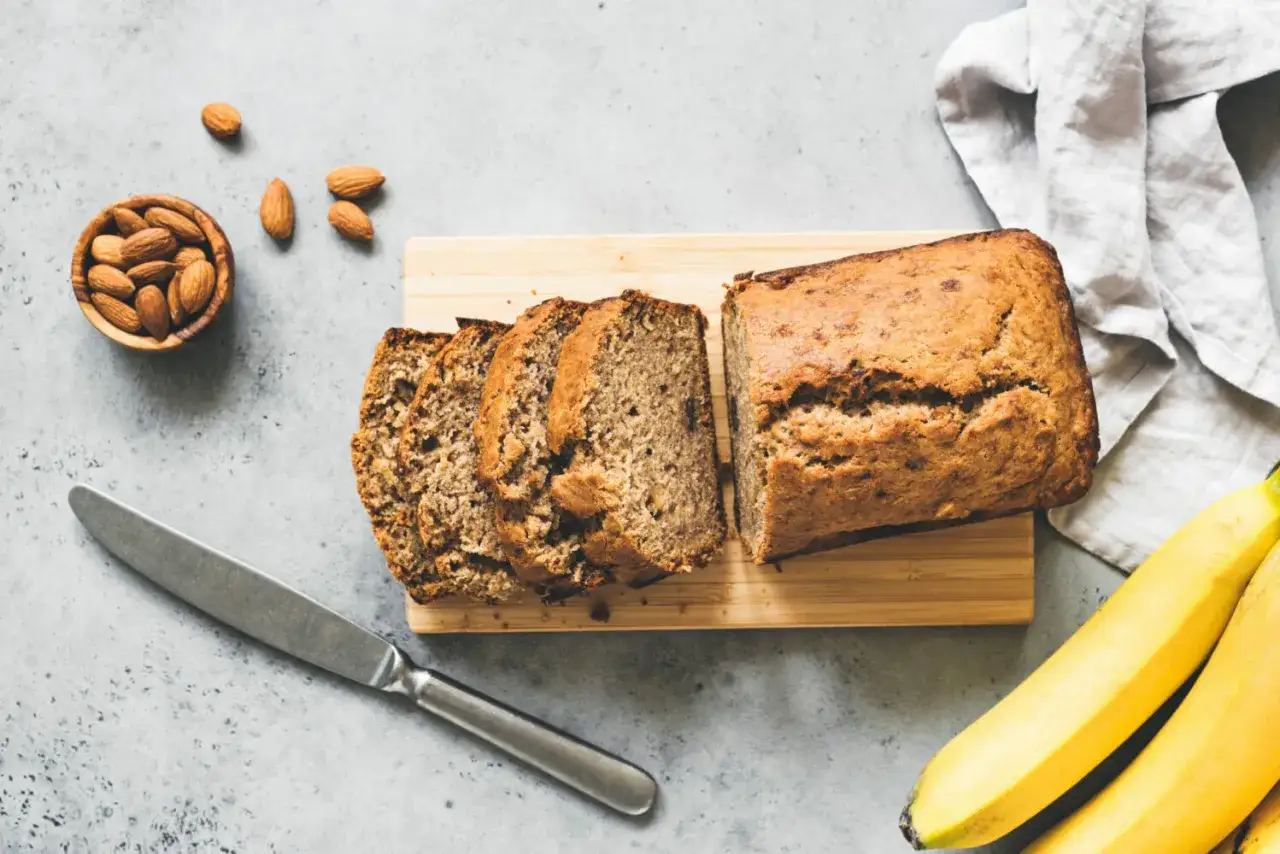 Tranche de banana bread healthy sur planche en bois, amandes et bananes à proximité.