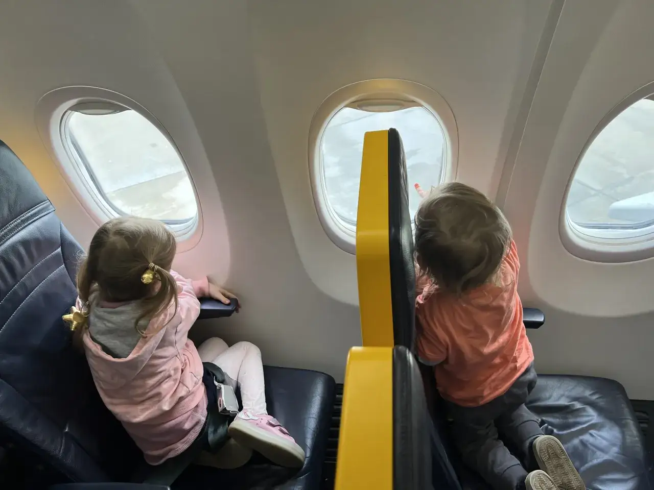 Dzieci patrzą przez okno samolotu Ryanair. Dziewczynka w różowej bluzie i chłopiec w pomarańczowej koszulce.
