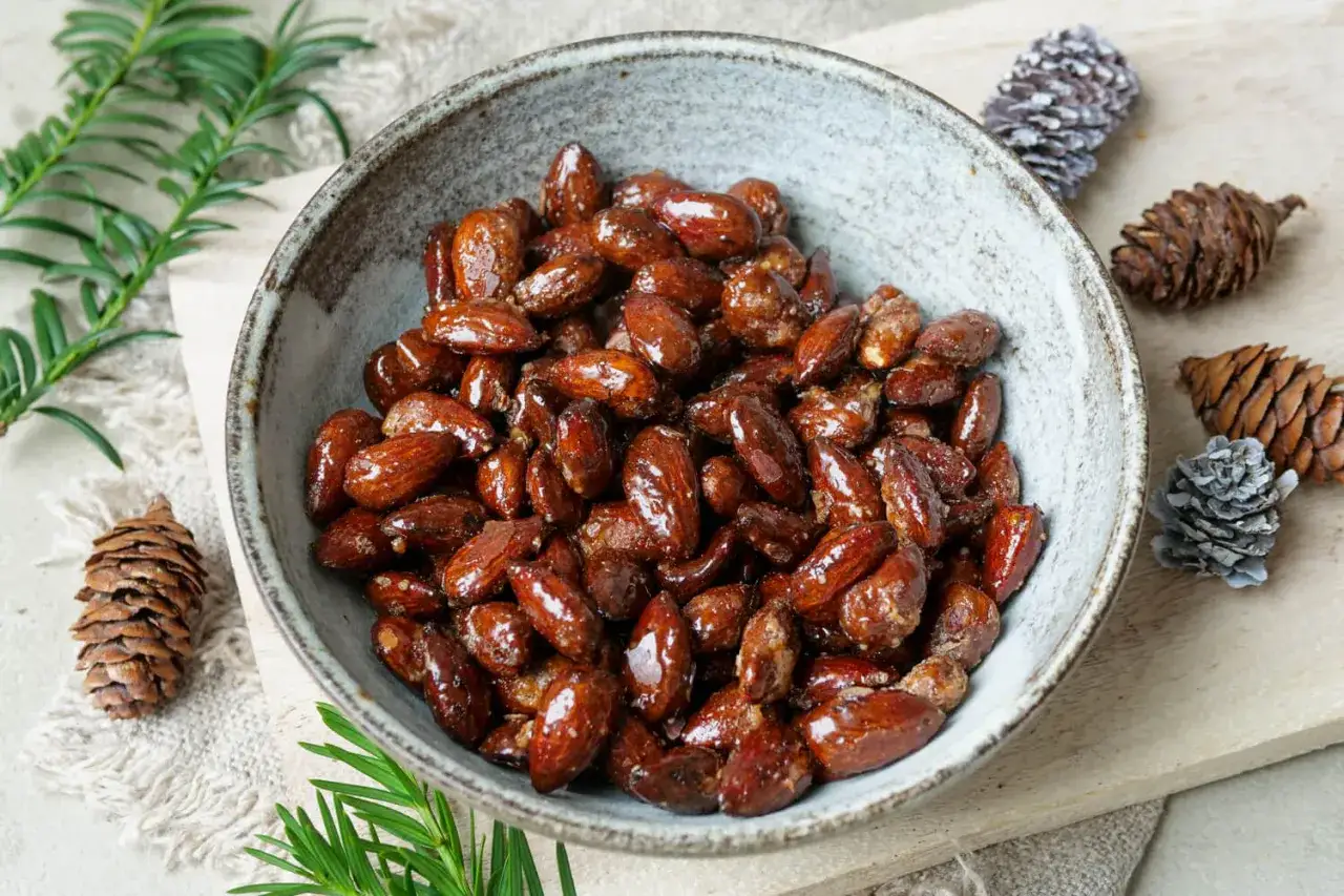 Schüssel mit glasierten gebrannten Mandeln, perfekt für den Airfryer. Weihnachtliche Tannenzapfen und Tannenzweige im Hintergrund.