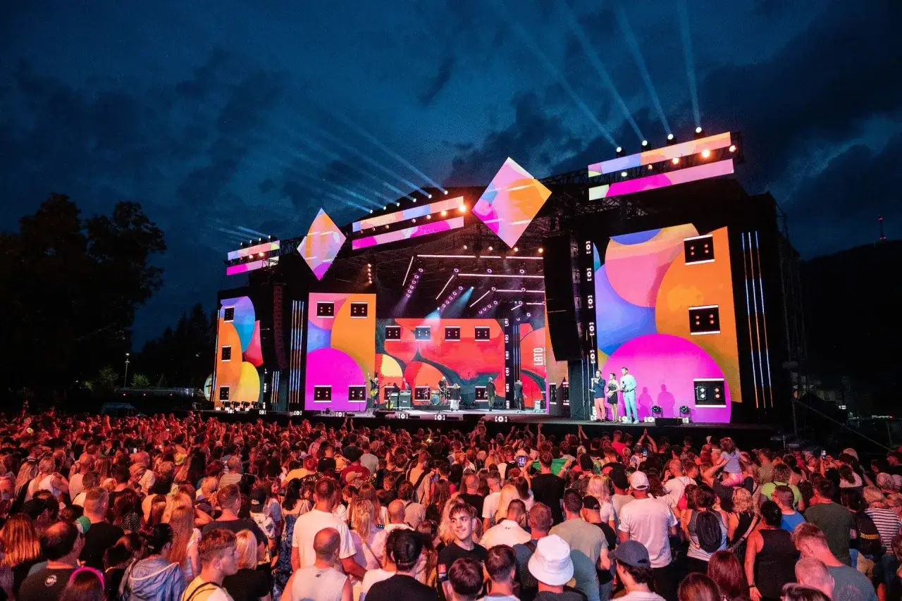 Koncert na żywo, tłum ludzi pod sceną, gdzie artyści wykonują utwory Stachursky'ego. Kolorowe światła i wizualizacje na ekranach.