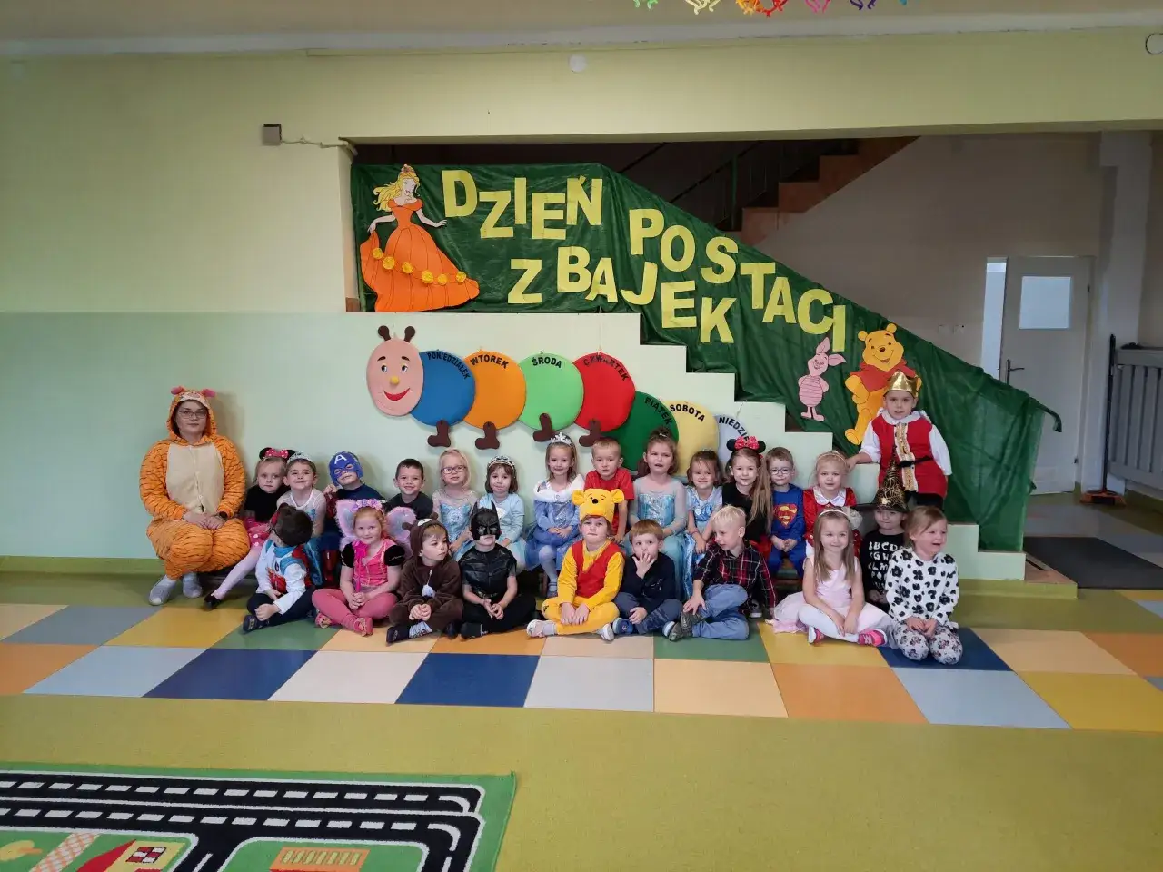 Dzień postaci z bajek w przedszkolu. Dzieci przebrane za bohaterów bajek, od księżniczek po superbohaterów, tworzą barwny korowód.