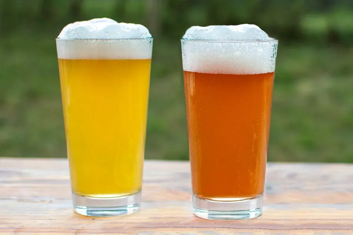 Deux verres de bière, l'un jaune pâle, l'autre ambré, sur une table en bois. Une pale ale ou une IPA pour se rafraîchir.