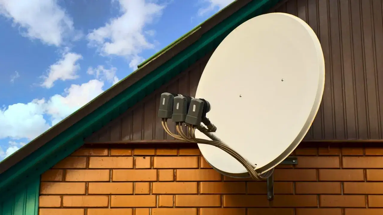 Duży biały talerz do satelity zamontowany na ceglanej ścianie domu, pod zielonym okapem dachu.