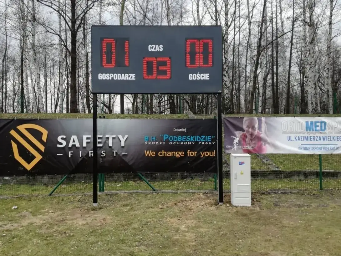 Wynik polska: gospodarze 01, goście 03. Tablica wyników na boisku, za nią drzewa i banery reklamowe.