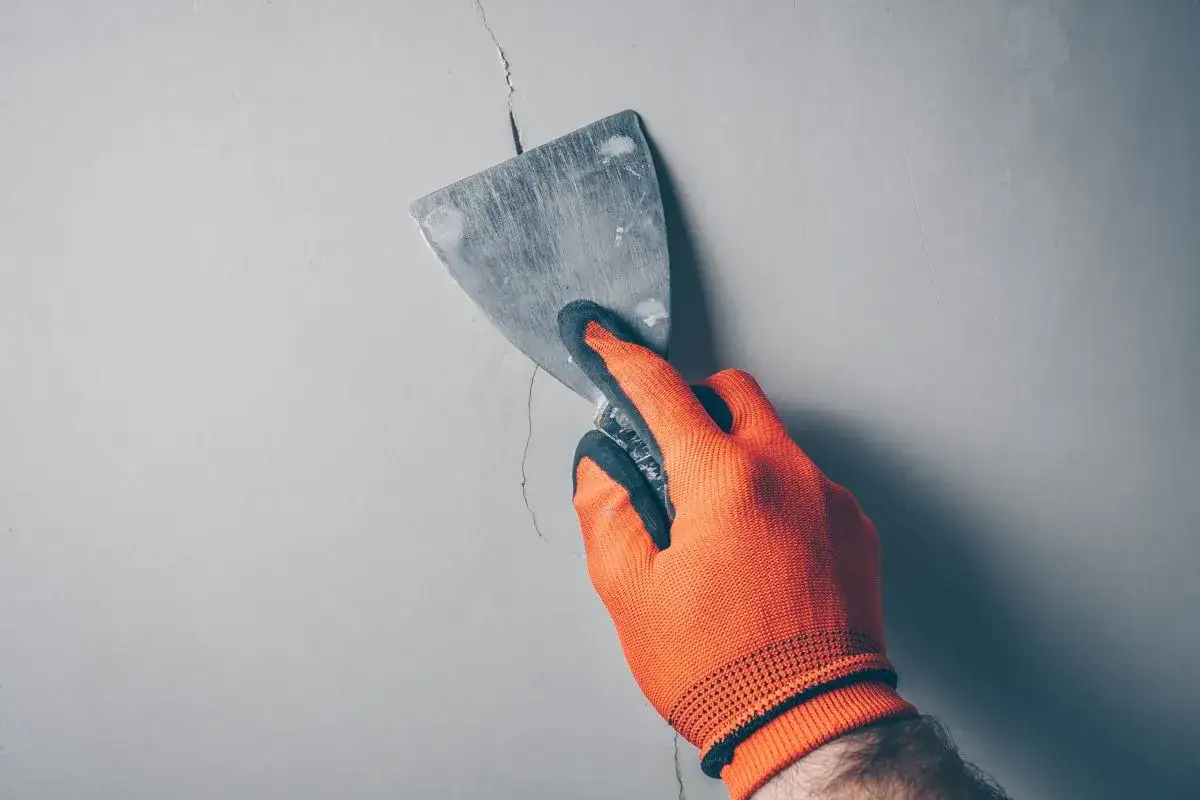 Ręka w pomarańczowej rękawicy zacierająca pęknięcie w ścianie szpachlą. Idealne rozwiązanie, czym wypełnić głębokie pęknięcia w ścianie.