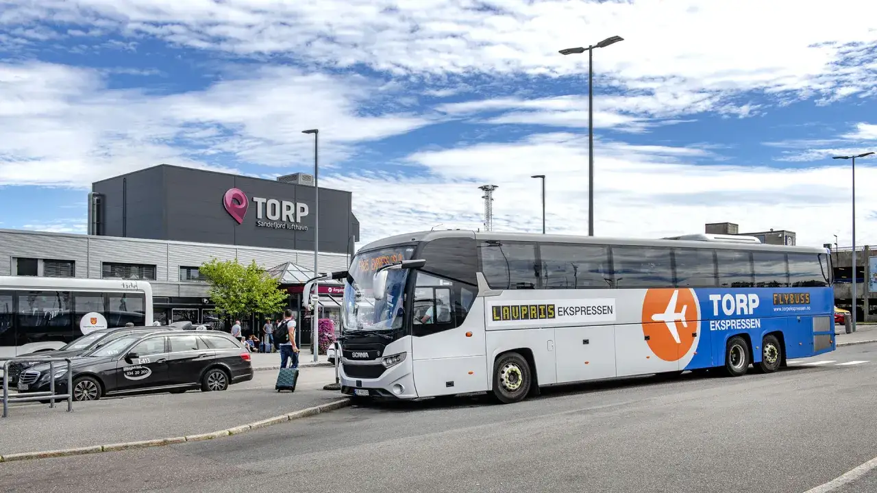 Autobus Flybuss Torp Ekspressen czeka na pasażer&oacute;w przy lotnisku Oslo Torp. Obok stoi czarny samoch&oacute;d.