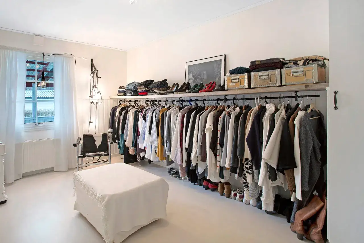 Ett stilrent, litet walk in closet med kl&auml;der p&aring; galgar, skor p&aring; golvet och f&ouml;rvaringsl&aring;dor p&aring; hyllan. En vit ottoman st&aring;r framf&ouml;r en f&aring;t&ouml;lj.