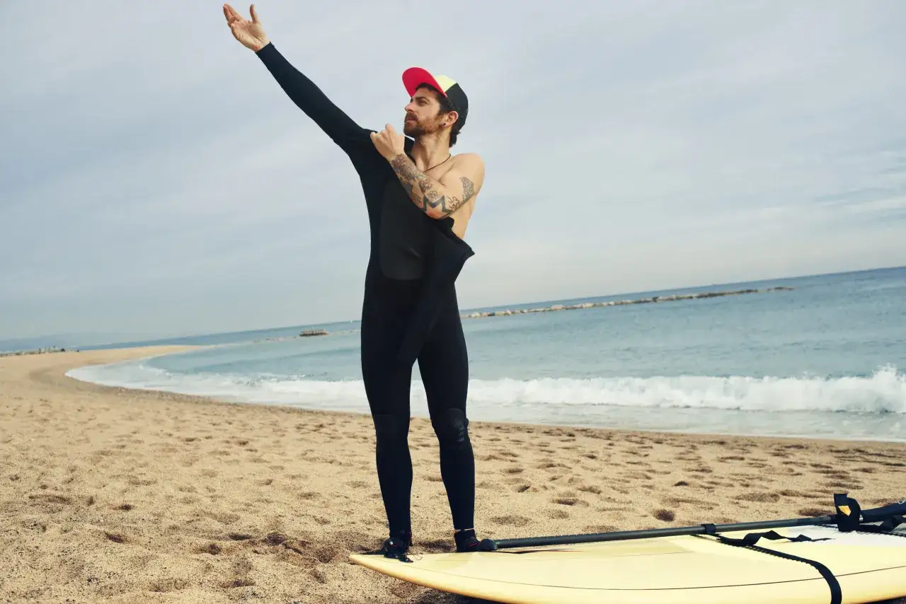 Mężczyzna z tatuażami zdejmuje piankę do pływania na plaży, obok deski SUP.