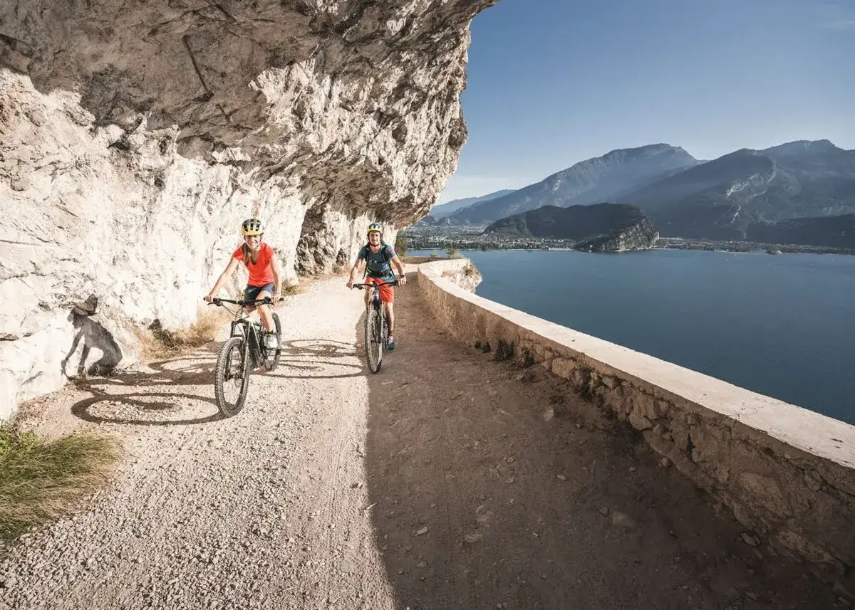 Zwei Radfahrer genießen eine **E-Bike Tour am Gardasee** auf einem schmalen Weg entlang einer Felswand mit Blick auf den See und die Berge.