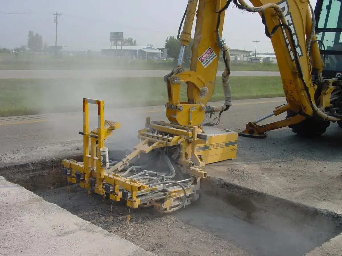 Une foreuse jaune perc&eacute;e le b&eacute;ton d'une route, soulevant de la poussi&egrave;re.