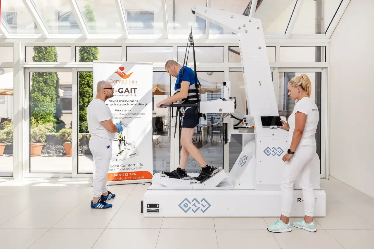 Rehabilitacja pacjenta z niedowładem nóg na urządzeniu Gait Trainer. Fizjoterapeuci wspierają proces nauki chodu.