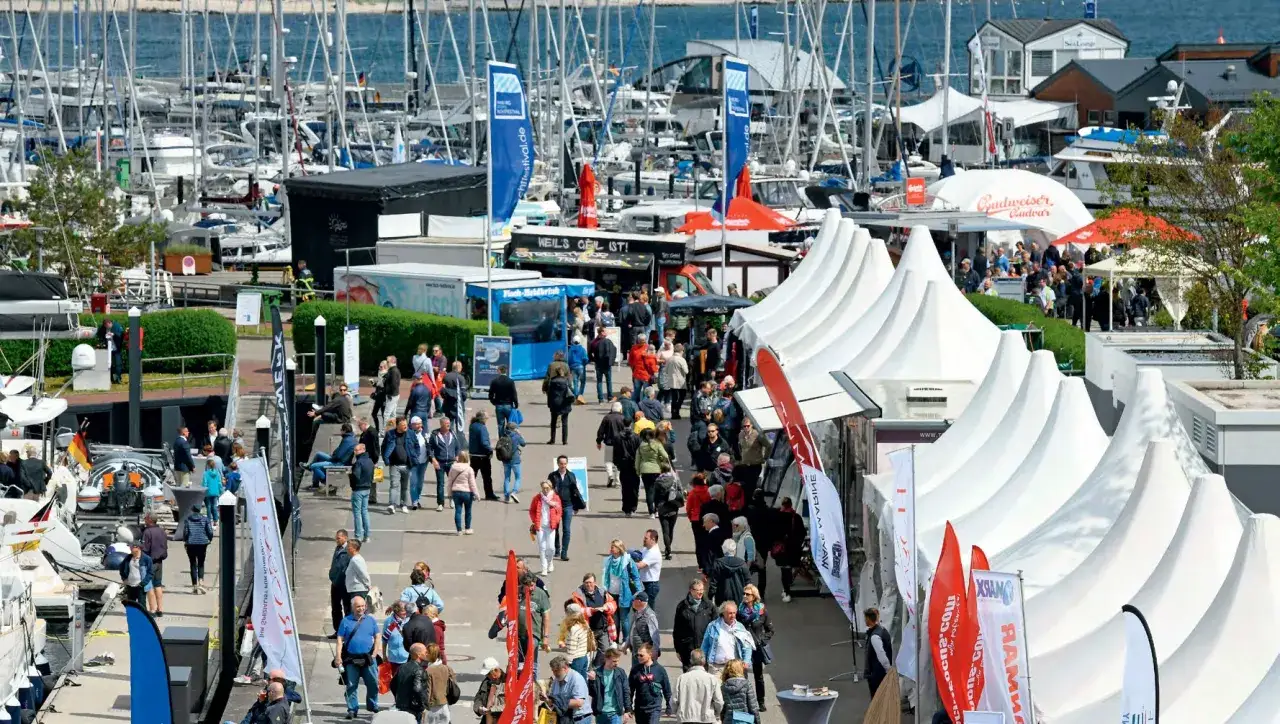 Besucher bummeln &uuml;ber die ancora boat show, vorbei an wei&szlig;en Zelten und vielen Segelbooten im Hafen.
