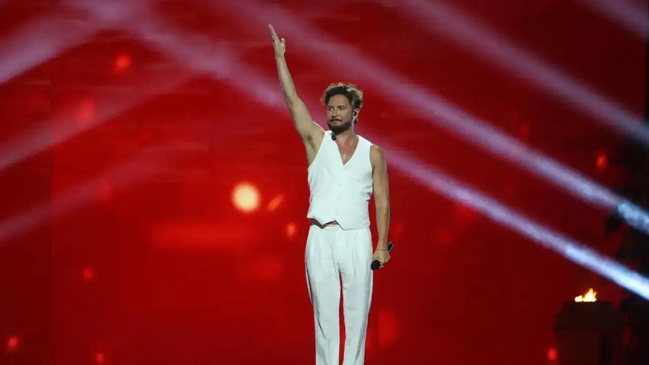 Manuel Carrasco saluda al público en su gira, con un traje blanco y luces rojas de fondo.