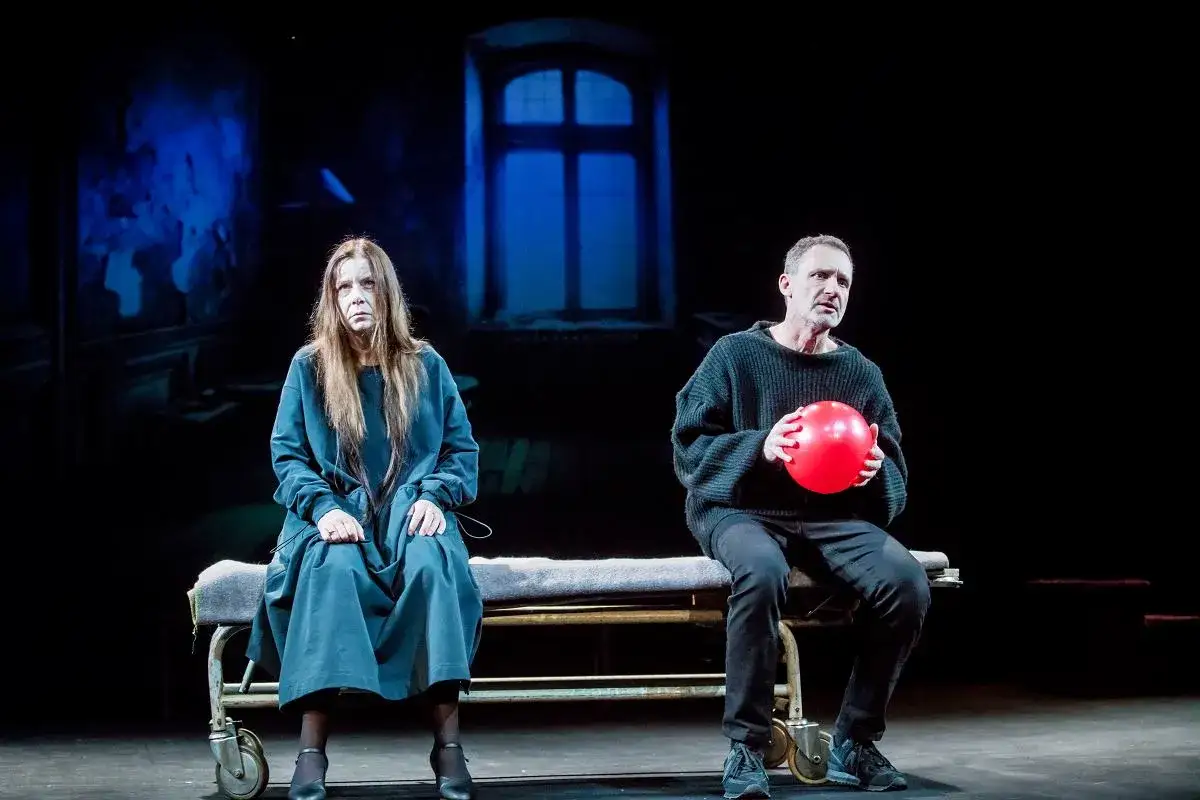 Agata Kulesza w teatrze, siedzi obok mężczyzny z czerwoną piłką. Mroczna scenografia.