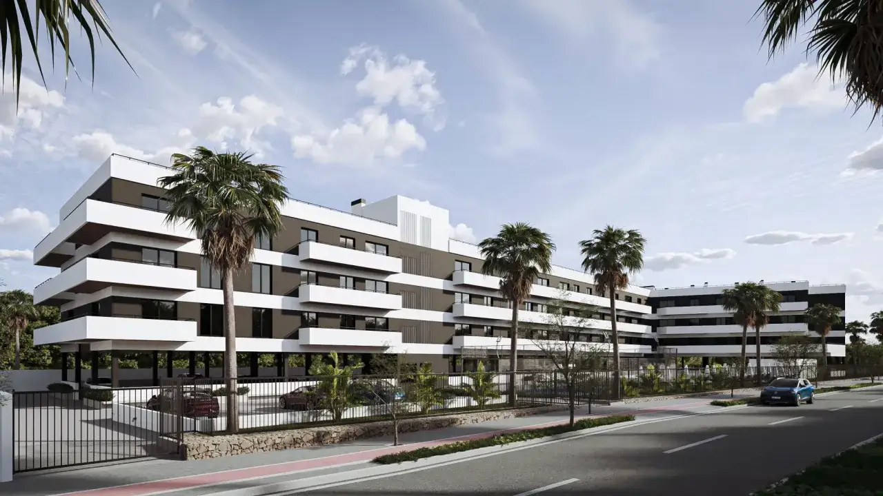 Edificio moderno con balcones blancos y fachada oscura, rodeado de palmeras. Un coche azul circula por la calle. Son viviendas VPO en Alicante.