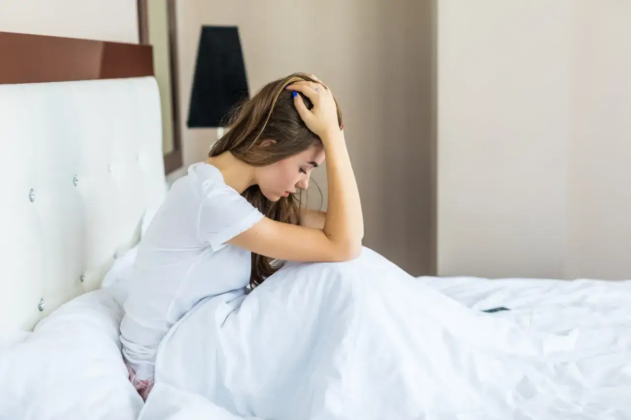 Mujer joven en la cama, con la cabeza entre las manos, sintiendo ansiedad al despertar.