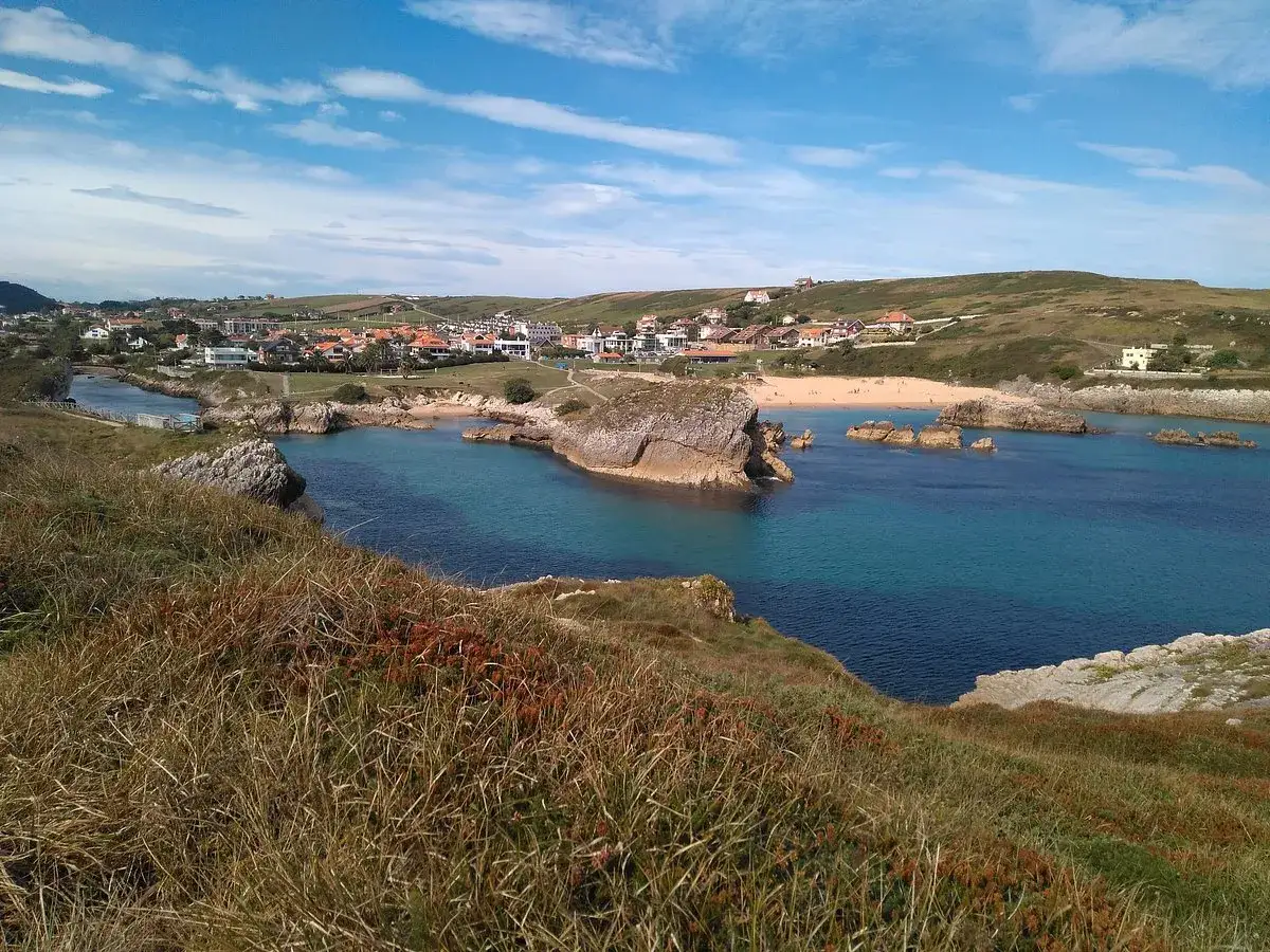 Vista de la playa San Juan de la Canal con aguas turquesas, rocas y un pueblo costero al fondo.