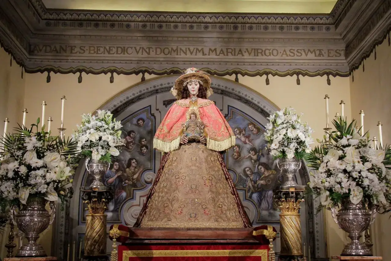 Virgen del Roc&iacute;o Pastora, vestida con traje dorado y manto rosa, sostiene al Ni&ntilde;o Jes&uacute;s. Flores blancas y &aacute;ngeles la rodean.