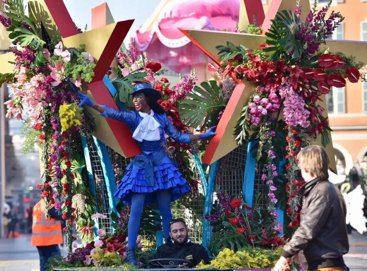 Una mujer con un traje azul y sombrero baila en una carroza floral del carnaval de Niza.